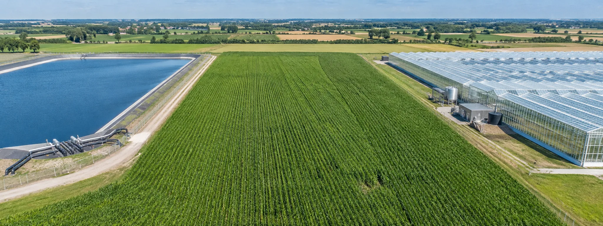 Aerial view of a water reservoir, a corn field and a greenhouse side by side